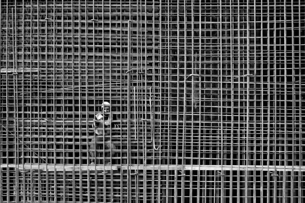 Monochrome image of a worker behind steel reinforcement bars in a construction site.