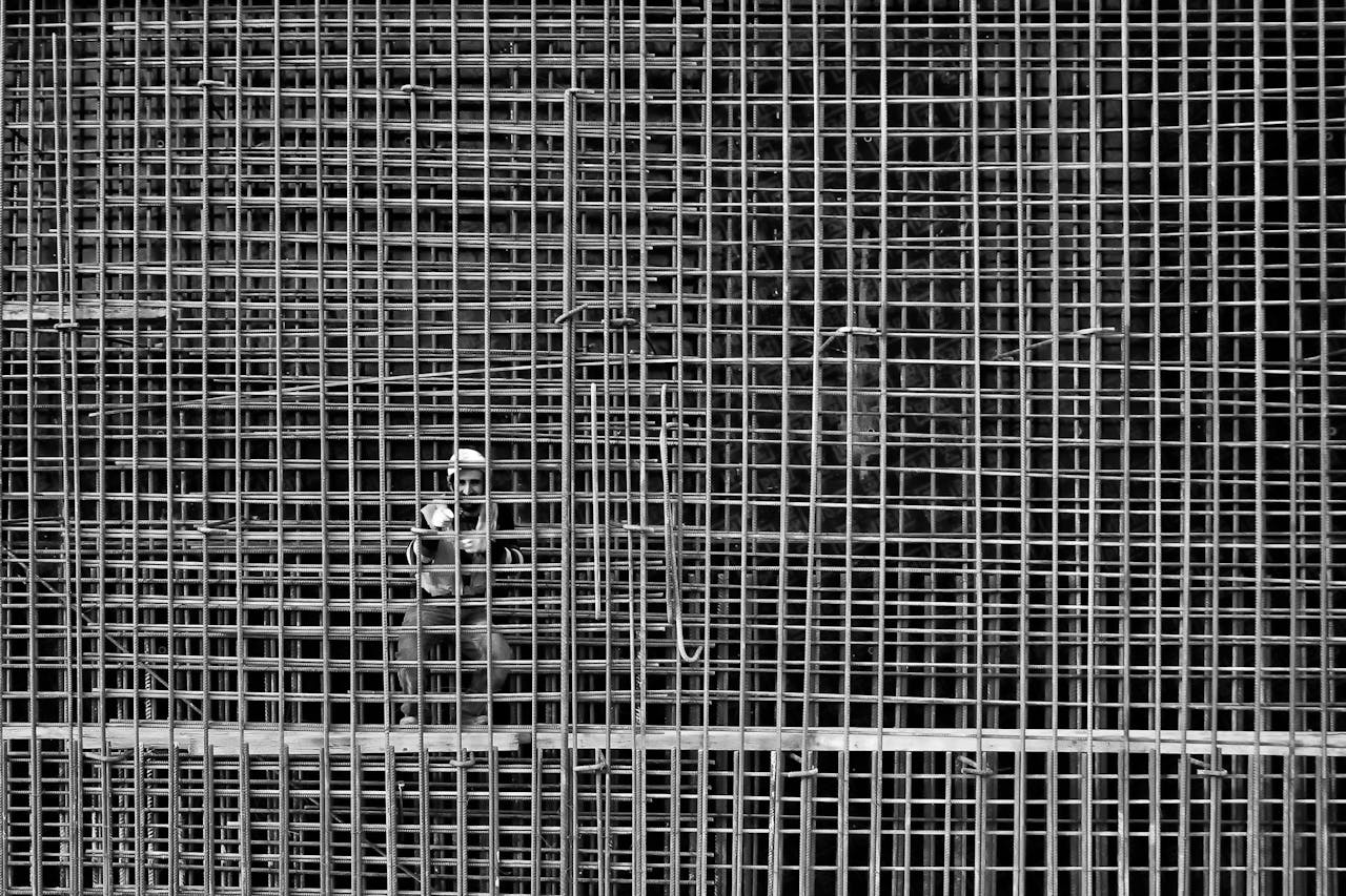 Monochrome image of a worker behind steel reinforcement bars in a construction site.